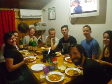 Um grupo de oito amigos se senta ao redor de uma mesa de madeira, compartilhando uma refeição e bebidas, sorrindo, com uma TV na parede e um ambiente aconchegante.