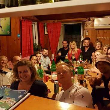 Um grupo de amigos se reúne em torno de uma mesa de madeira em um ambiente interno, sorrindo e saboreando bebidas. Garrafas e copos cobrem a mesa, criando uma atmosfera animada e aconchegante.