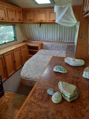 Essa imagem mostra o interior de um trailer ou RV, com uma cama com mosquiteiro, armários de madeira, uma janela, algumas pedras pintadas e uma bancada.
