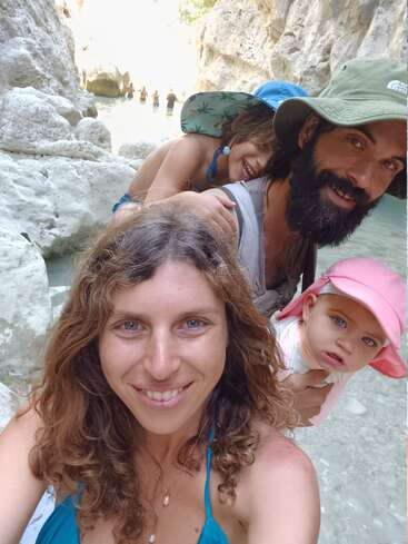 Uma família de quatro pessoas sorri perto de águas cristalinas entre penhascos rochosos. A mãe tira uma selfie, registrando momentos felizes com seu parceiro, filho e filha bebê.