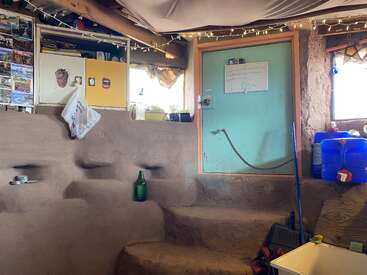 Dieser gemütliche, rustikale Raum verfügt über eine irdene Treppe, Regale mit Postkarten, eine blaue Tür mit einem Whiteboard, Wasserbehälter, Lichterketten und einfache Aufbewahrung.