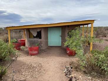 Ein kleines, rustikales Haus mit einer türkisfarbenen Tür und einem gelben Dach steht in einer kargen Landschaft, umgeben von roten Pflanzgefäßen und spärlicher Wüstenvegetation unter einem wolkenverhangenen Himmel.