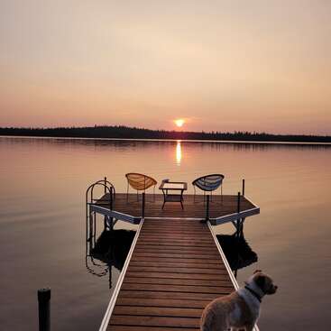 A dog stands on a wooden dock overlooking a calm lake at sunset. Two chairs and a table invite relaxation, creating a peaceful, serene atmosphere.