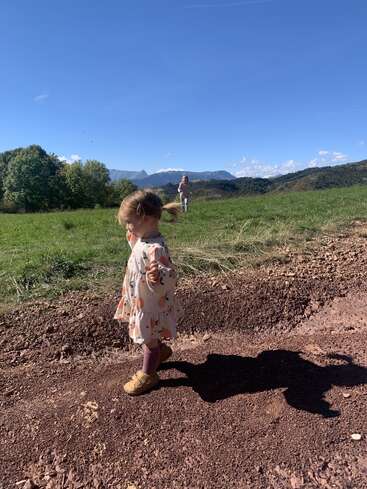 Ein junges Mädchen in einem gemusterten Kleid läuft auf einem Feldweg, während ein anderes Kind weiter hinten auf einer Wiese unter einem klaren blauen Himmel steht.