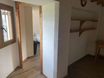 Dieses Bild zeigt einen kleinen, einfachen Innenraum mit weißen Wänden, Holzverkleidung, offenen Regalen, einem Holztisch, einem Fenster und einer Tür, die zu einem Schlafzimmer führt.