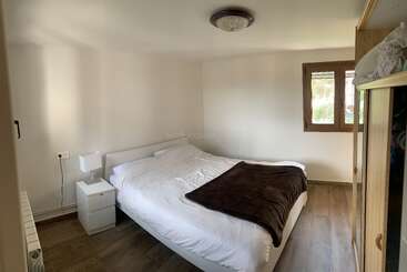 Dies ist ein kleines, minimalistisches Schlafzimmer mit weißen Wänden, einem Doppelbett, einer braunen Decke, einem Beistelltisch, einer Lampe, einem Kleiderschrank, Holzböden und einem Fenster. Sehr gemütlich.