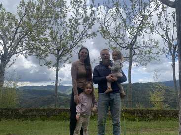 Una familia de cuatro miembros está al aire libre, rodeada de árboles. Detrás de ellos, las montañas y el cielo nublado crean un telón de fondo natural y tranquilo. Los padres sonríen con sus dos hijos.