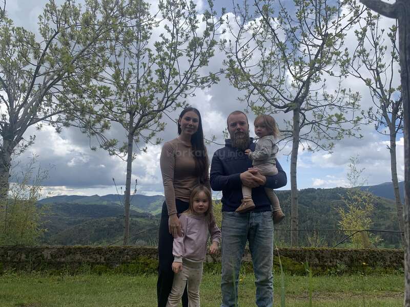 Eine vierköpfige Familie steht im Freien, umgeben von Bäumen. Hinter ihnen bilden die malerischen Berge und der bewölkte Himmel eine friedliche, natürliche Kulisse. Die Eltern lächeln mit ihren beiden Kindern.