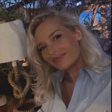 Une femme aux cheveux blonds et à la chemise bleu clair sourit doucement à l'appareil photo. Elle est à l'intérieur, avec des plantes et une lampe à côté d'elle.