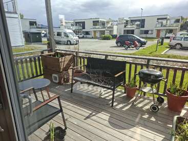 The image shows a cozy wooden porch with a bench, round table, chairs, potted plants, a barbecue grill, and a view of modern apartment buildings outside.