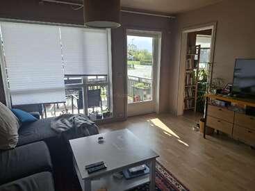A cozy living room with a grey couch, coffee table, TV, and bookshelf. Natural light enters through large windows and a door leading to a balcony.