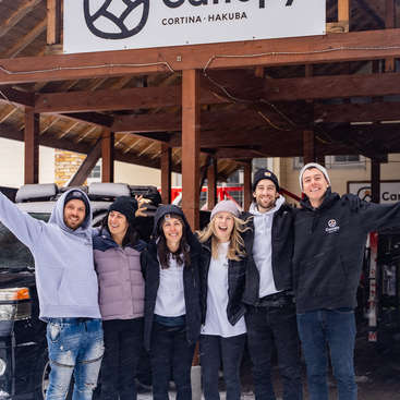 The image shows six people standing in front of a building with a sign that reads \"Canopy Cortina Hakuba\", wearing winter clothing and posing for a photo.