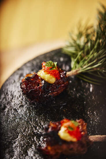 Dos trozos de carne carbonizada con hueso, aderezados con salsa cremosa, huevas rojas y hierbas, servidos en un plato negro texturizado junto a agujas de pino verdes.