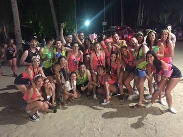 The image depicts a group of approximately 30 people posing for a photo on a sandy beach at night, with some holding drinks and buckets.