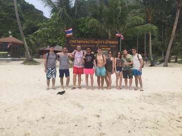 The image depicts a group of nine people standing on a sandy beach, posing for a photo in front of a sign that reads \"Mue Ang Thong National Park\" in Thai and English.