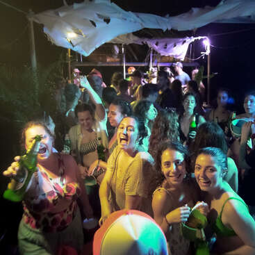 The image depicts a lively party scene with a group of young adults gathered together, holding drinks and smiling, surrounded by a festive atmosphere.