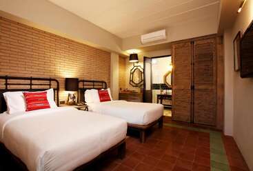 The image depicts a hotel room with a double bed, a desk, and a chair, featuring a large window and a door, with a white wall and a gray floor. The room is well-lit and has a neutral color scheme.