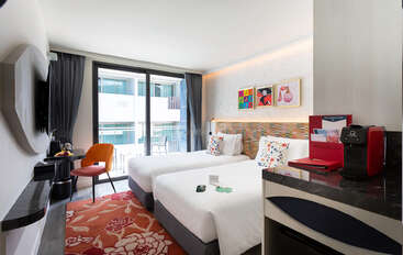 The image depicts a hotel room with a bed, desk, and chair, featuring a large window with a view of the city. The room is well-lit and has a modern aesthetic.