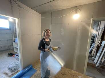 La imagen muestra a una mujer de pie en una habitación en construcción, sosteniendo una bandeja con herramientas, rodeada de materiales de construcción y herramientas, con una puerta y una ventana visibles al fondo.