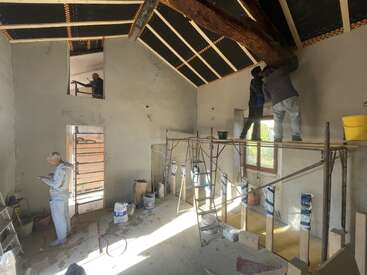 La imagen muestra una habitación en construcción, con un techo alto con vigas a la vista, andamios y obreros realizando tareas de renovación.