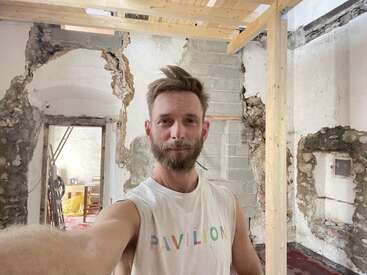 Un hombre de pelo corto y castaño se hace un selfie en una habitación en reformas, con las paredes vistas y un marco de madera, vestido con una camiseta de tirantes blanca con la palabra "PAVILION".