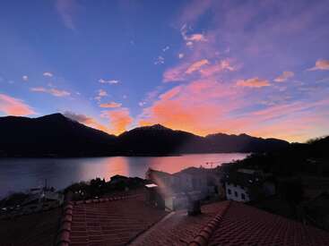 La imagen representa una serena puesta de sol sobre un lago, con montañas al fondo y los tejados de una ciudad en primer plano, sobre un cielo vibrante.