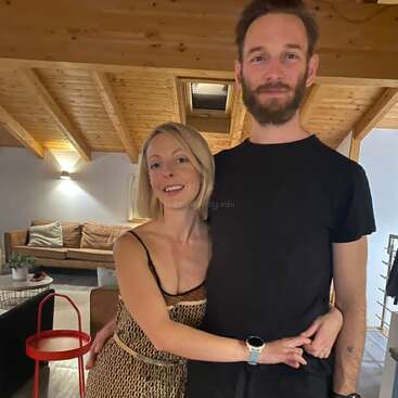 La imagen muestra a una pareja de pie en un acogedor salón, abrazados, con la mujer a la izquierda y el hombre a la derecha, ambos sonriendo a la cámara.