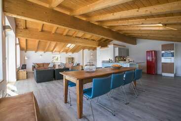 La imagen muestra una habitación espaciosa, de techo alto, con una gran mesa y sillas, y bien iluminada, lo que sugiere un comedor formal o una sala de estar. Las paredes son blancas y el suelo de madera.