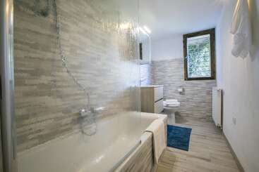La imagen muestra un cuarto de baño con bañera, lavabo e inodoro, en una combinación de colores blanco y gris, con una ventana y una puerta, y una pared blanca al fondo.