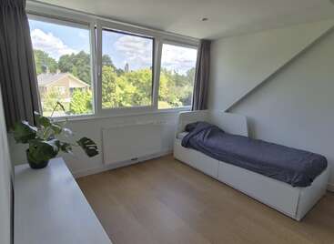 Il s'agit d'une chambre à coucher minimaliste avec un lit simple, de grandes fenêtres, un sol en bois clair, des murs blancs, des rideaux gris et une plante en pot sur la commode.