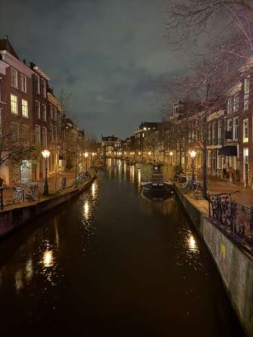 Scène de nuit sereine sur un canal dans une ville européenne, avec des maisons en briques, des lampadaires, des vélos garés, des bateaux et une eau calme reflétant des lumières chaudes et dorées. Atmosphère paisible.