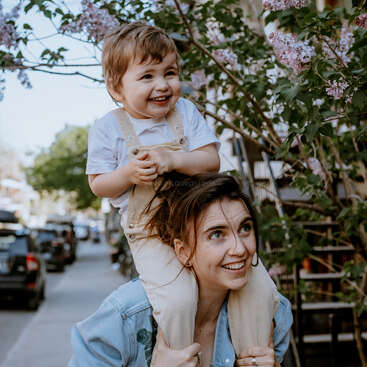A woman with dark hair carries a toddler on her shoulders, both smiling, in front of a flowering tree and a city street with parked cars.