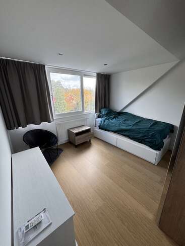 Um quarto pequeno e minimalista com piso de madeira, paredes brancas, uma cama de solteiro com roupa de cama escura, uma cadeira preta, mesa de cabeceira, cômoda e uma grande janela com cortinas.