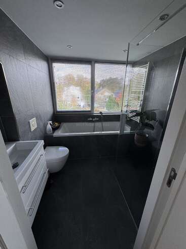 Esse banheiro moderno tem azulejos escuros, uma pia branca elegante, vaso sanitário montado na parede, banheira com janelas grandes com persianas, uma divisória de vidro para o chuveiro e um vaso de plantas.