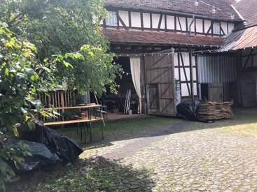 Ein rustikaler Innenhof mit Kopfsteinpflaster, Grünpflanzen und einer offenen Scheune mit Holztüren. Gestapelte Tische, Bänke und Holzstapel schaffen eine gemütliche, ländliche Atmosphäre.