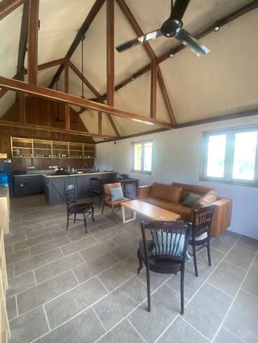This image shows a spacious kitchen and dining area with high vaulted ceilings, exposed wooden beams, modern furniture, a brown sofa, and ample natural light from windows.