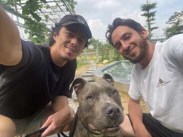 Two smiling men take a selfie outdoors with a gray dog. They look happy. Behind them is a small pond, greenery, and a partly cloudy sky.
