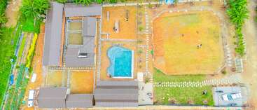 This aerial image shows a compound with several buildings, a uniquely shaped swimming pool, a grassy field, a parking area with cars, surrounded by greenery.