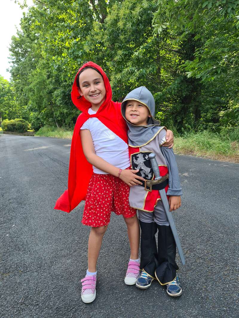 Das Bild zeigt zwei Kinder, die auf einer Straße stehen. Das eine trägt einen roten Umhang und das andere ist als Ritter verkleidet und posiert für ein Foto.