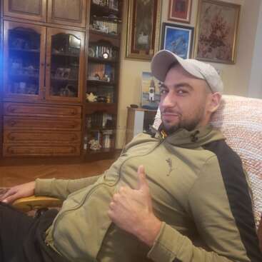 A man wearing a cap and beige jacket relaxes on a chair, smiling and giving a thumbs up, with a cozy living room background behind him.