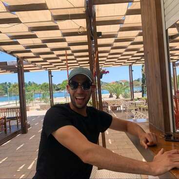 A man wearing sunglasses and a cap is smiling at an outdoor beachside cafe. Sunlight filters through a wooden pergola. Palm trees and water are visible.