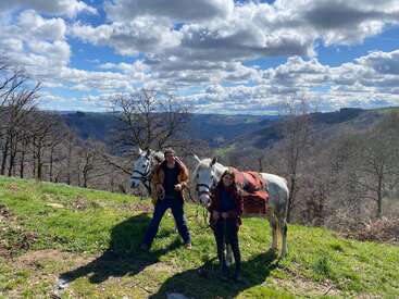 A man and woman stand in a scenic, mountainous landscape with two white horses. Leafless trees, green grass, and dramatic clouds enhance the natural beauty around them.