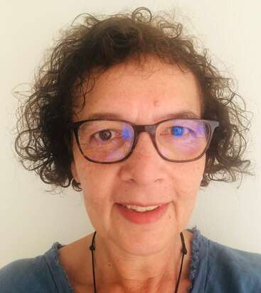 Das Bild zeigt eine Frau mit lockigen braunen Haaren, die eine schwarz gerahmte Brille und ein blaues Hemd trägt, vor einem weißen Hintergrund.