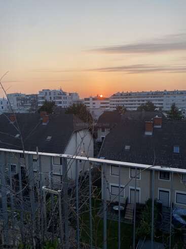 Das Foto zeigt einen ruhigen Sonnenuntergang über den Dächern von Wohnhäusern und Apartmentgebäuden, mit kahlen Ästen im Vordergrund und sanften Orange- und Rosatönen am Himmel.