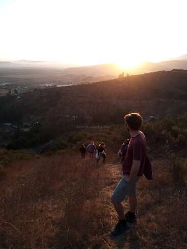 La imagen representa a un hombre de pie en la ladera de una colina, contemplando una puesta de sol sobre un valle, con tres personas caminando detrás de él en la distancia.