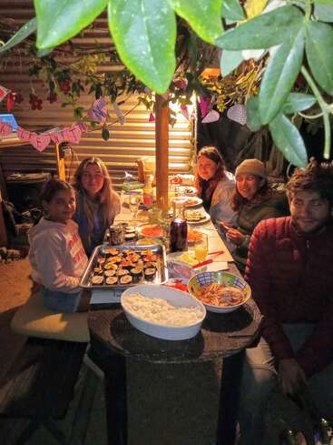 La imagen muestra a un grupo de cinco personas sentadas a una larga mesa cargada con un surtido de comida y bebida, rodeadas de un ambiente festivo.