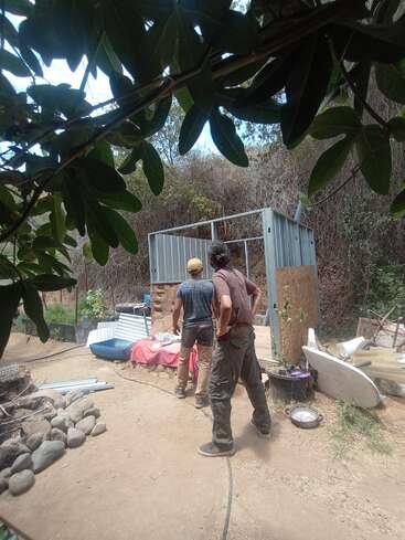 La imagen muestra a dos hombres de pie en un patio, rodeados de materiales de construcción y escombros, con una estructura metálica al fondo y árboles en primer plano.