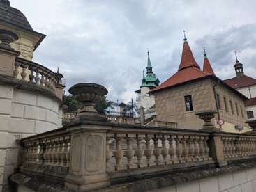 Historic stone balustrades and urns frame the scene, with an old building featuring red-tiled rooftops and church spires under a cloudy, overcast sky. Peaceful atmosphere.