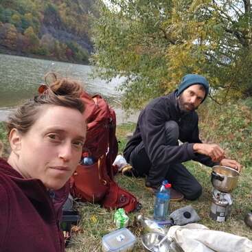 Two people are camping by a lake, surrounded by greenery. One person is cooking on a portable stove while the other takes a selfie. Outdoor gear scattered around.