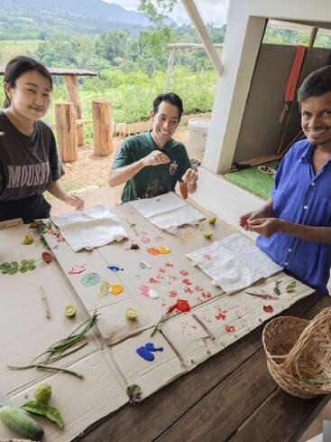 Trois personnes sourient et s'amusent à peindre des tissus en plein air. La table est recouverte de peinture colorée, de feuilles, de légumes et de matériaux naturels, entourée d'un paysage magnifique.